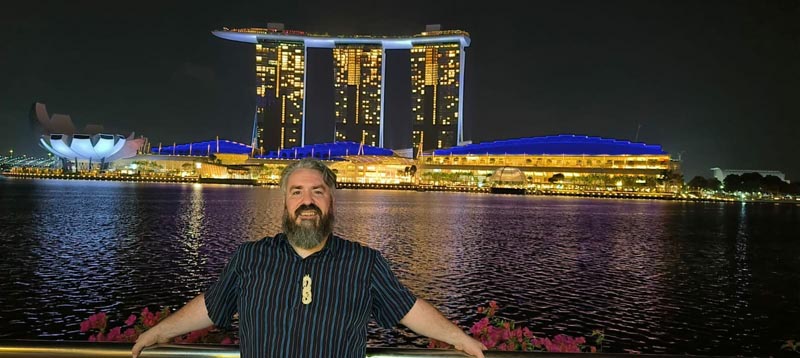 Neil Wallace in front of a large building in Singapore in the evening when lights feature across the harbour